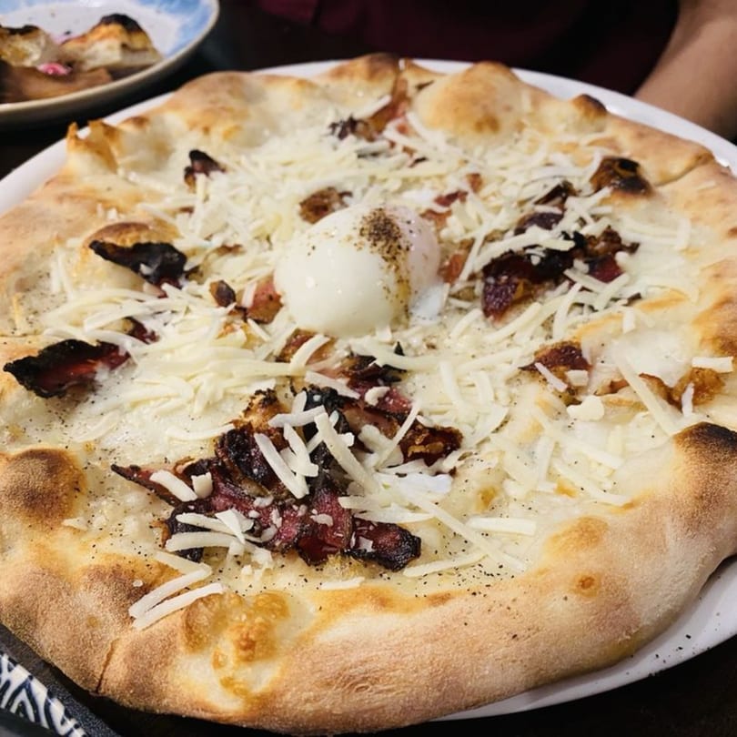 A white pizza with bacon, shredded cheese, and a poached egg on a white plate.