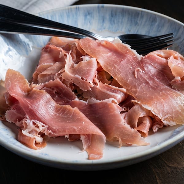 Thinly sliced prosciutto with white fat marbling, served on a light blue and white plate with cutlery.